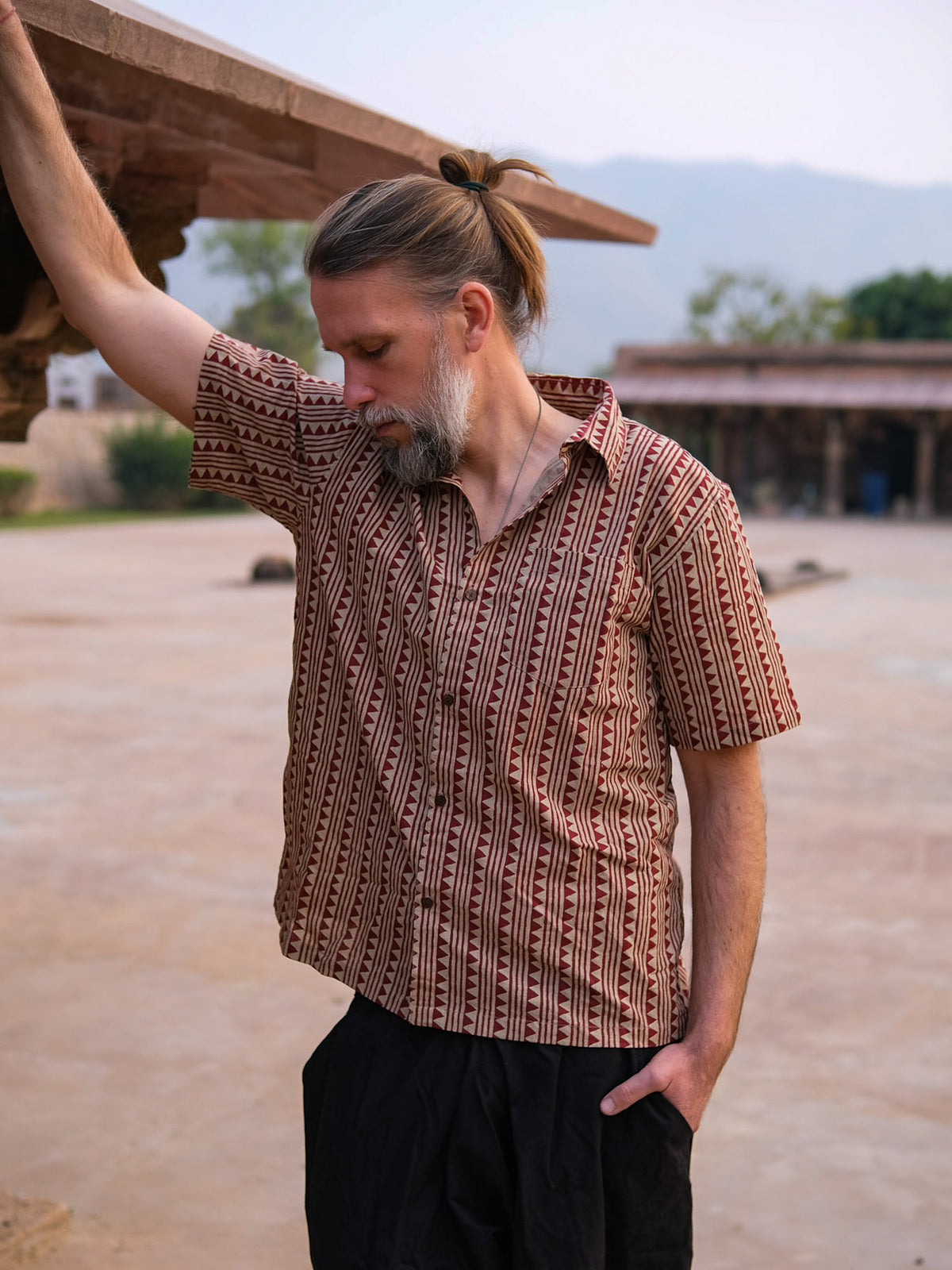 A man with a beard and tied-back hair stands outdoors, leaning against a structure with one hand while looking down. He wears the Tribal Triangle shirt—a unisex piece showcasing a distinct Rajasthan print—paired with dark pants. In the background, distant buildings and trees are visible.