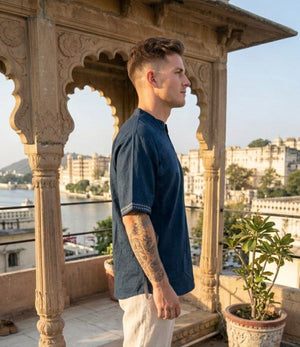 A man wearing the Midnight Nomad kurta stands in profile on a balcony with ornate arches, overlooking a river and historic buildings under clear skies.