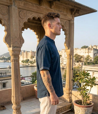 A man wearing the Midnight Nomad kurta stands in profile on a balcony with ornate arches, overlooking a river and historic buildings under clear skies.