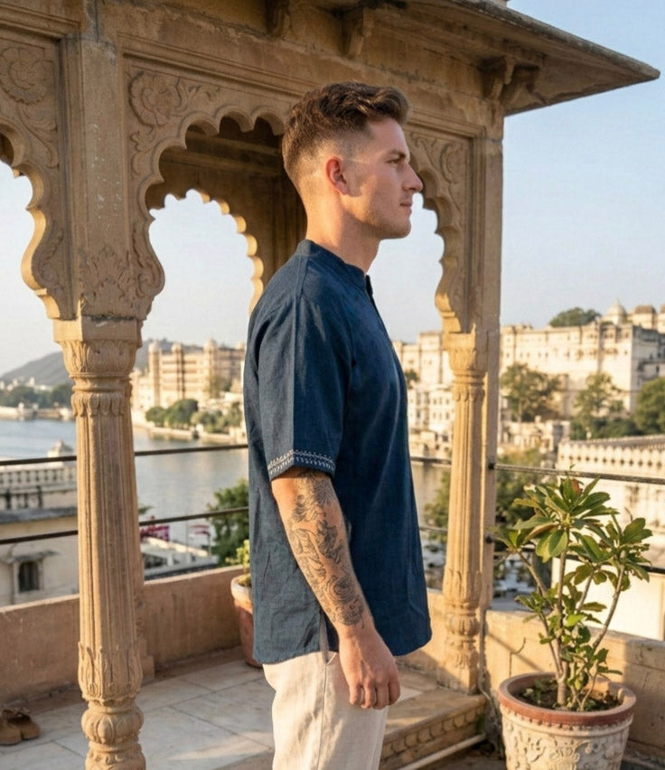 A man wearing the Midnight Nomad kurta stands in profile on a balcony with ornate arches, overlooking a river and historic buildings under clear skies.