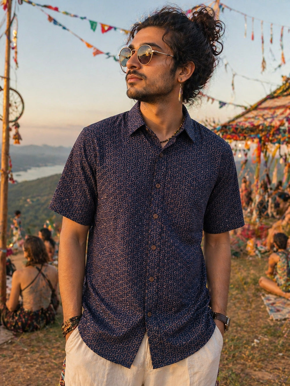 A man wearing the Neel shirt and sunglasses stands outdoors at a festival, with colorful decorations and people sitting on the grass in the background.