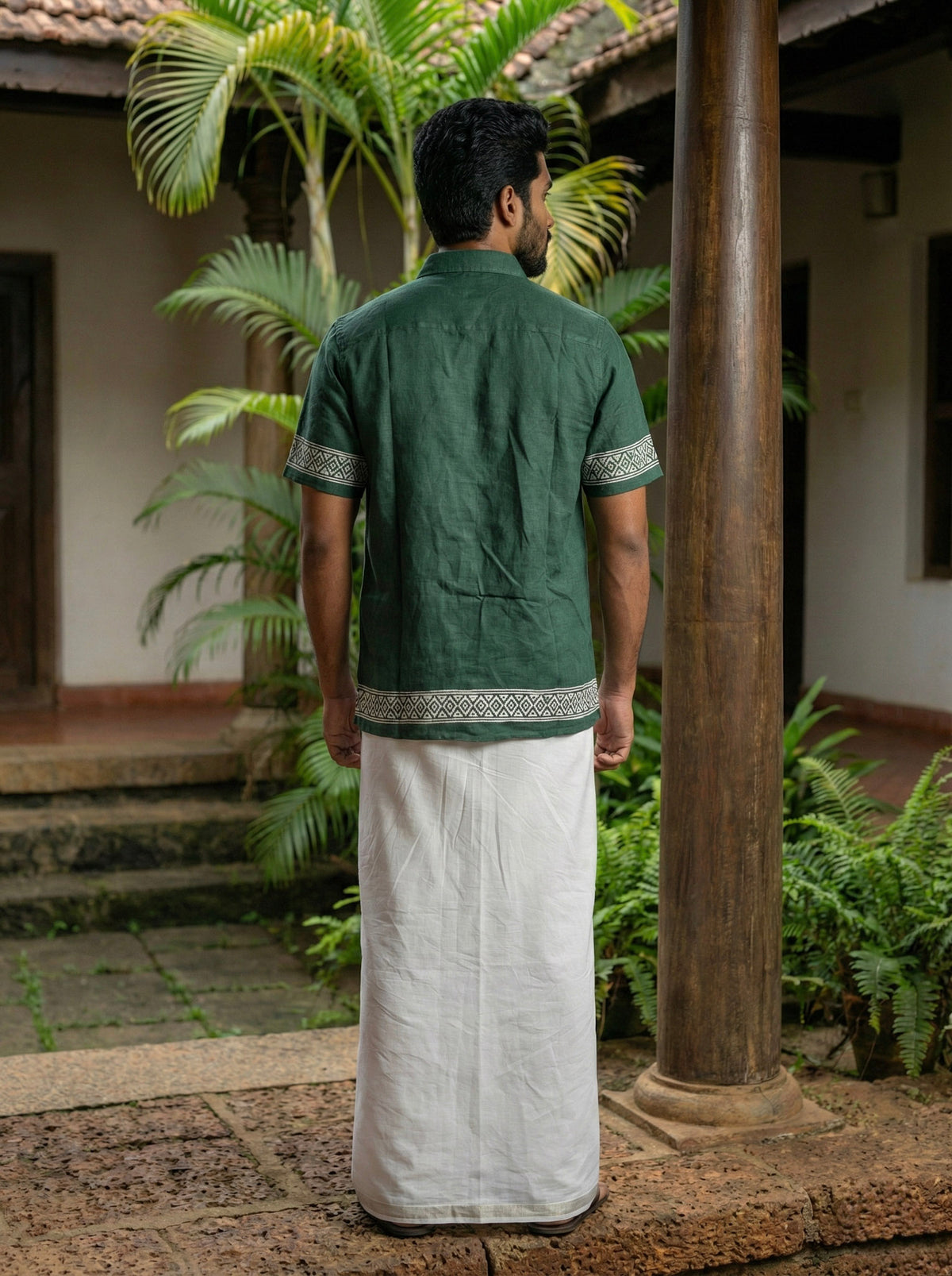 A man stands with his back to the camera in an outdoor courtyard filled with tropical plants, wearing the Adhir Shirt—crafted from green cotton with patterned trim—and a white traditional dhoti.