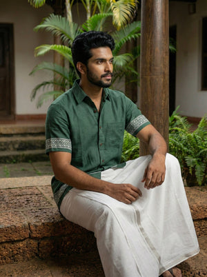 A man with a beard sits outdoors on a low stone wall, surrounded by greenery and a wooden pillar, wearing the Adhir Shirt—a contemporary green cotton piece with patterned cuffs—paired with a traditional white dhoti.