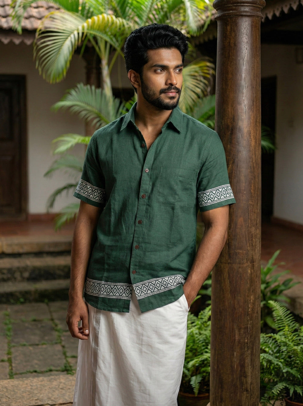 A man wearing the Adhir Shirt, an ethnic contemporary green short-sleeved cotton shirt with handcrafted-inspired patterns on the sleeves and hem, paired with a white dhoti, stands beside a wooden pillar in a courtyard featuring lush plants and traditional architecture in the background.