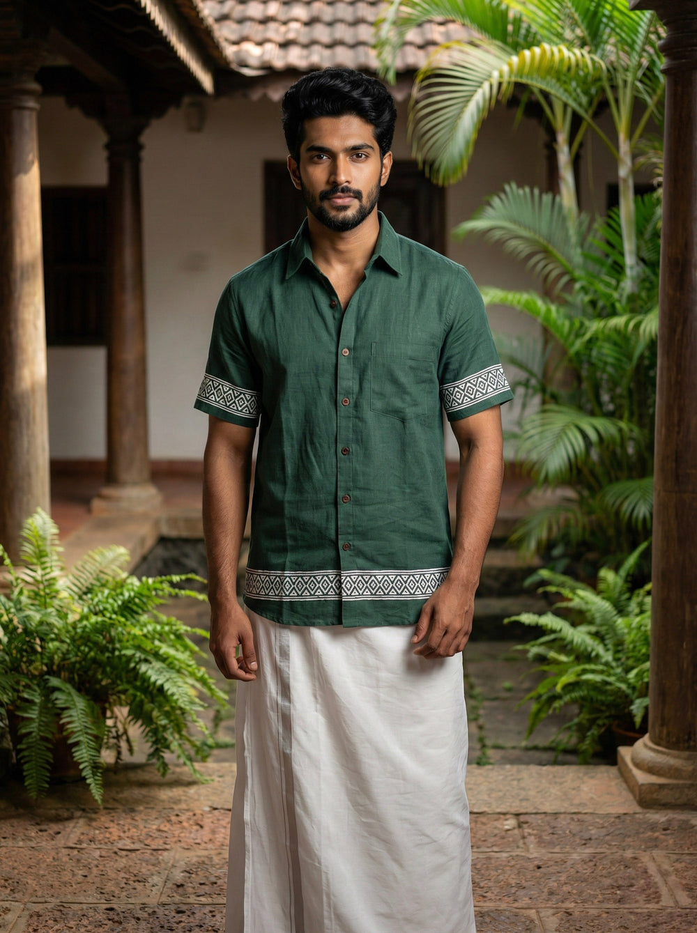 A man wearing the Adhir Shirt in green with patterned borders and a white dhoti stands in a traditional courtyard featuring plants and wooden pillars.