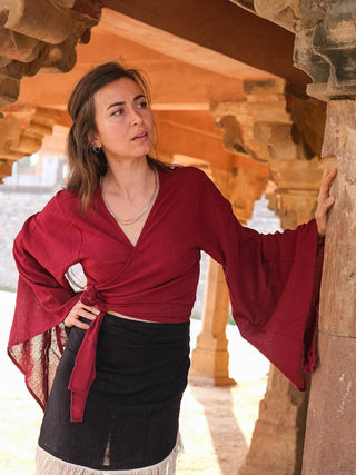 Wearing a Nidra Wrap top in red, a woman stands under a stone archway, leaning against a pillar. Her wide bell sleeves flow as she gazes aside, layered necklaces glinting. Sunlight filters through the arches, adding a boho charm to the scene with her black skirt.