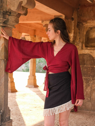 A woman in a red Nidra Wrap Top and a black fringed skirt stands inside a stone structure with arched openings. She leans against a pillar, gazing to the side as sunlight illuminates the scene.