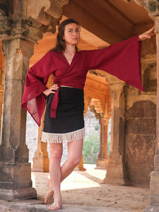 In a historic building with stone columns and arches, a barefoot woman poses with one arm raised. She wears the Nidra Wrap top—a boho-inspired burgundy piece with bell sleeves—and a black skirt with a fringe hem. Sunlight filters through the structure.