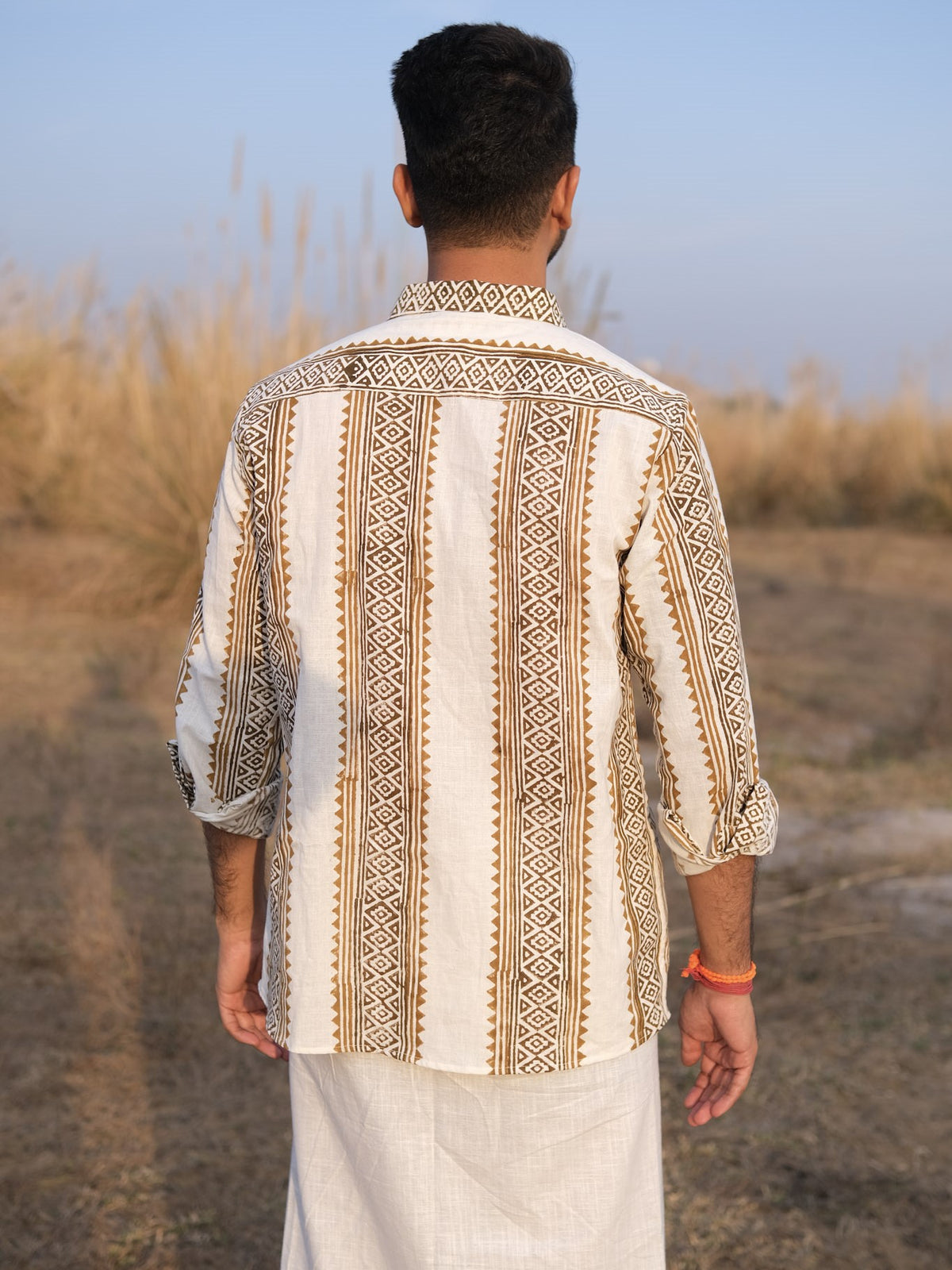 A man with short dark hair, wearing the Dwara Shirt—a block print cotton shirt in cream and brown—and white pants, stands outdoors on a dirt path, facing away from the camera with dry grass in the background.