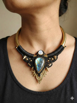 Dressed in a black top and the Elvara Necklace—a handmade macramé statement piece featuring gold metal, black thread, a round stone, and an oval stone with striking blue-green iridescence—a person perfectly captures the essence of boho tribal jewelry.