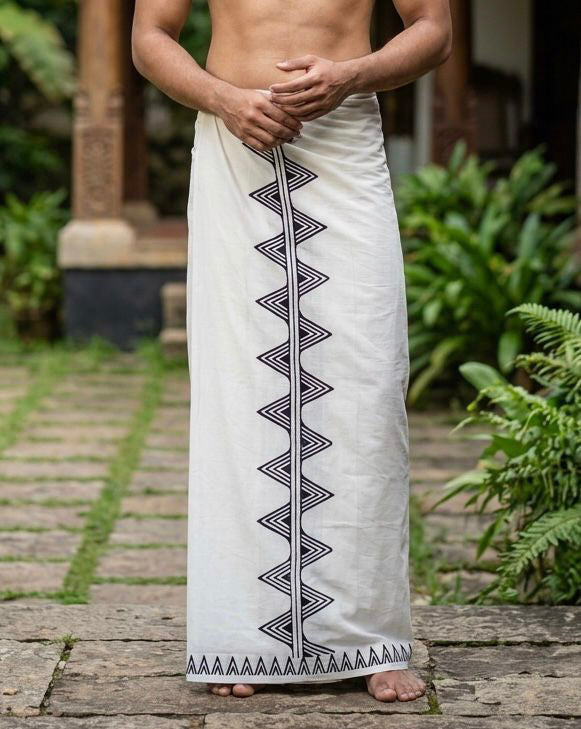 A person wearing the Sutra Dhoti, a traditional white cotton garment adorned with black geometric patterns crafted through hand block printing, stands barefoot on a stone path surrounded by greenery.