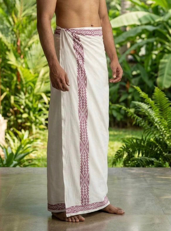 A man wearing the Elara Dhoti, featuring a geometric border, stands barefoot on a tiled floor with green plants in the background.