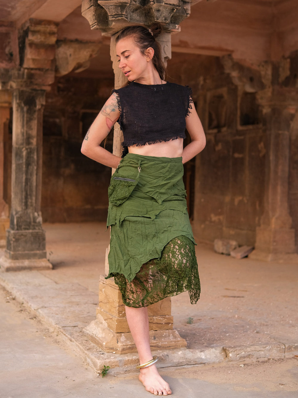 A person with a tattoo poses barefoot in ancient ruins, wearing a black fringed crop top and the Dhora Skirt—an asymmetrical green skirt featuring lace details and a detachable pocket—looking down with hands behind their back.