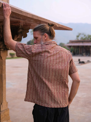 A man with a ponytail, seen from behind and dressed in the Tribal Triangle shirt with a vibrant Rajasthan-inspired print and black pants, leans against a stone column in an outdoor courtyard.
