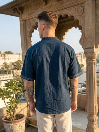 A man with tattoos, seen from behind, wears the Midnight Nomad kurta in a contemporary style paired with light pants, standing on a terrace with stone arches and potted plants, overlooking a city and water.