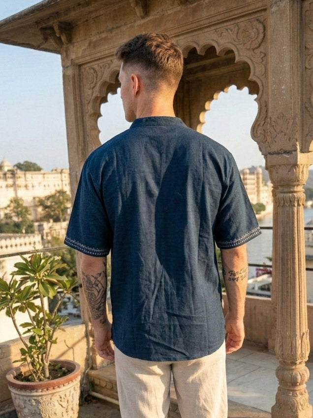 A man with tattoos, seen from behind, wears the Midnight Nomad kurta in a contemporary style paired with light pants, standing on a terrace with stone arches and potted plants, overlooking a city and water.