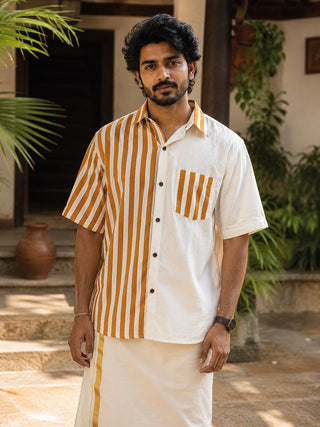 A man with dark hair and a beard wears a modern heritage Peetambar Shirt in natural color with yellow vertical stripes, paired with a matching cream dhoti featuring a subtle kasavu-gold stripe, as he stands outdoors near steps and greenery.