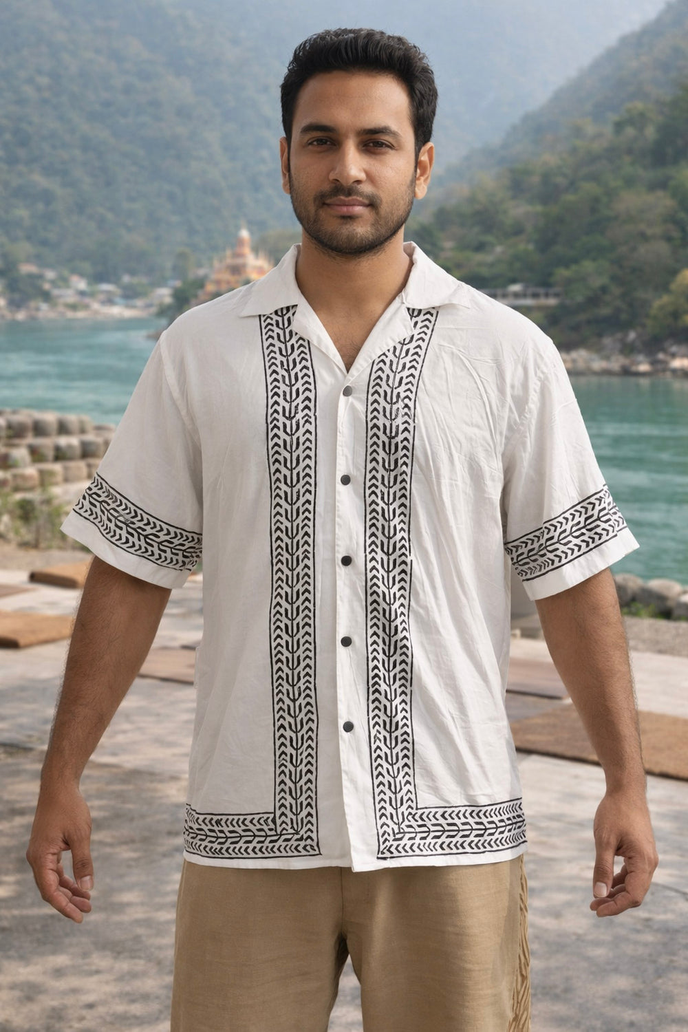 A man stands outdoors by a river, wearing the Zuri Shirt—a white short-sleeve hand block printed shirt with black patterns—paired with beige pants. Green hills and trees provide a scenic backdrop under a clear sky, highlighting the artisanal printing details of his resort-inspired Zuri Shirt.