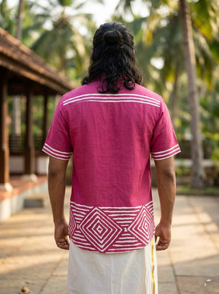 A person with long hair stands outdoors, facing away, wearing the Raktara Shirt—a berry-magenta geometric block-print shirt—paired with a white traditional garment, with palm trees and a tiled-roof building in the background.