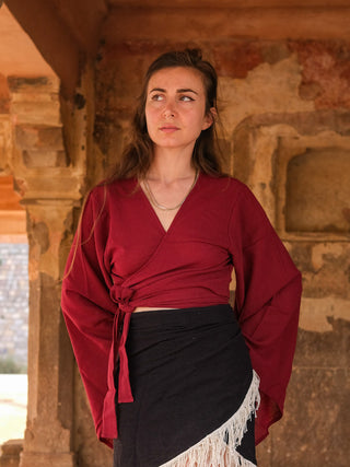 A woman with long hair stands before a rustic stone wall wearing the Nidra Wrap top featuring boho bell sleeves and a black skirt with white fringe, looking slightly to the side thoughtfully.