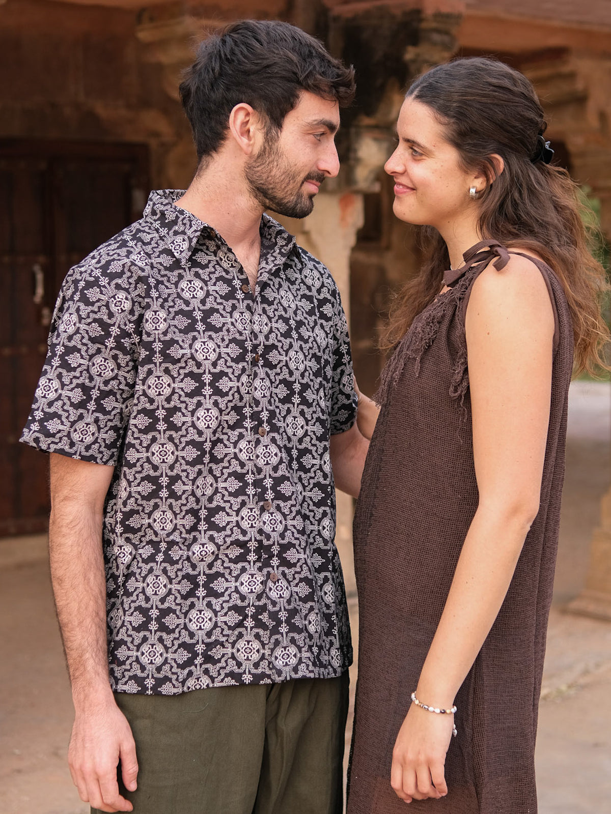 A man and woman stand close together, smiling at each other. The man is wearing a black Shipibo Shirt with short sleeves and green pants, while the woman wears a brown sleeveless dress. They are outdoors near a rustic building.