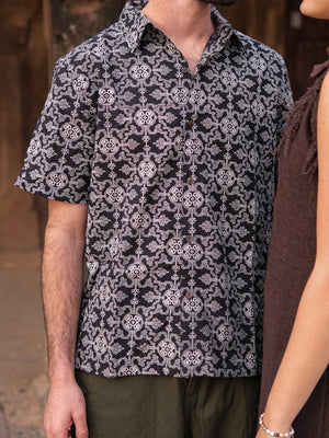 A man wearing the Shipibo Shirt—characterized by its short sleeves, black-and-white print, and lightweight cotton fabric—stands outdoors next to a woman in a sleeveless brown dress. Their upper bodies are visible from the shoulders down as they stand close together.