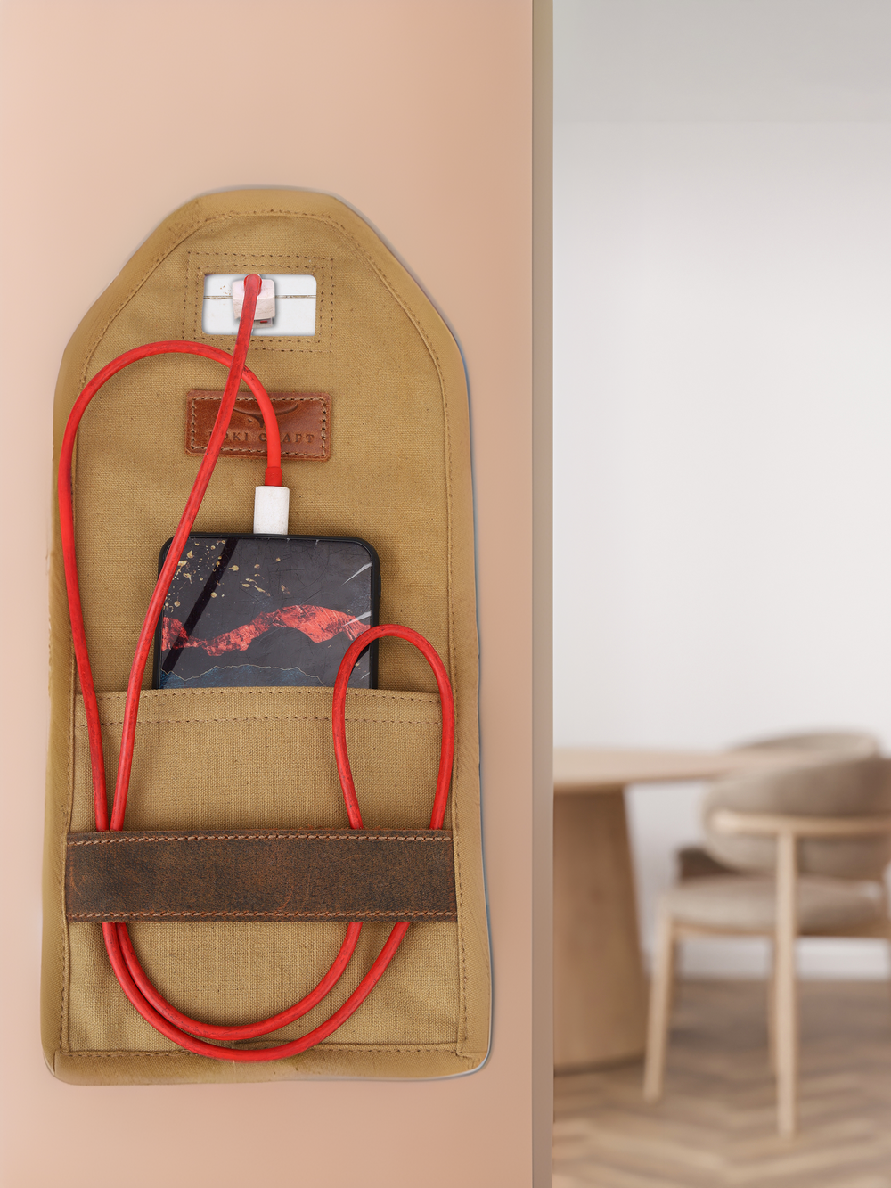The Energy Envelop, a brown wall-mounted fabric holder, securely charges your device with a red cable. In the background, a blurred wooden chair and table complete the minimalist room setting.