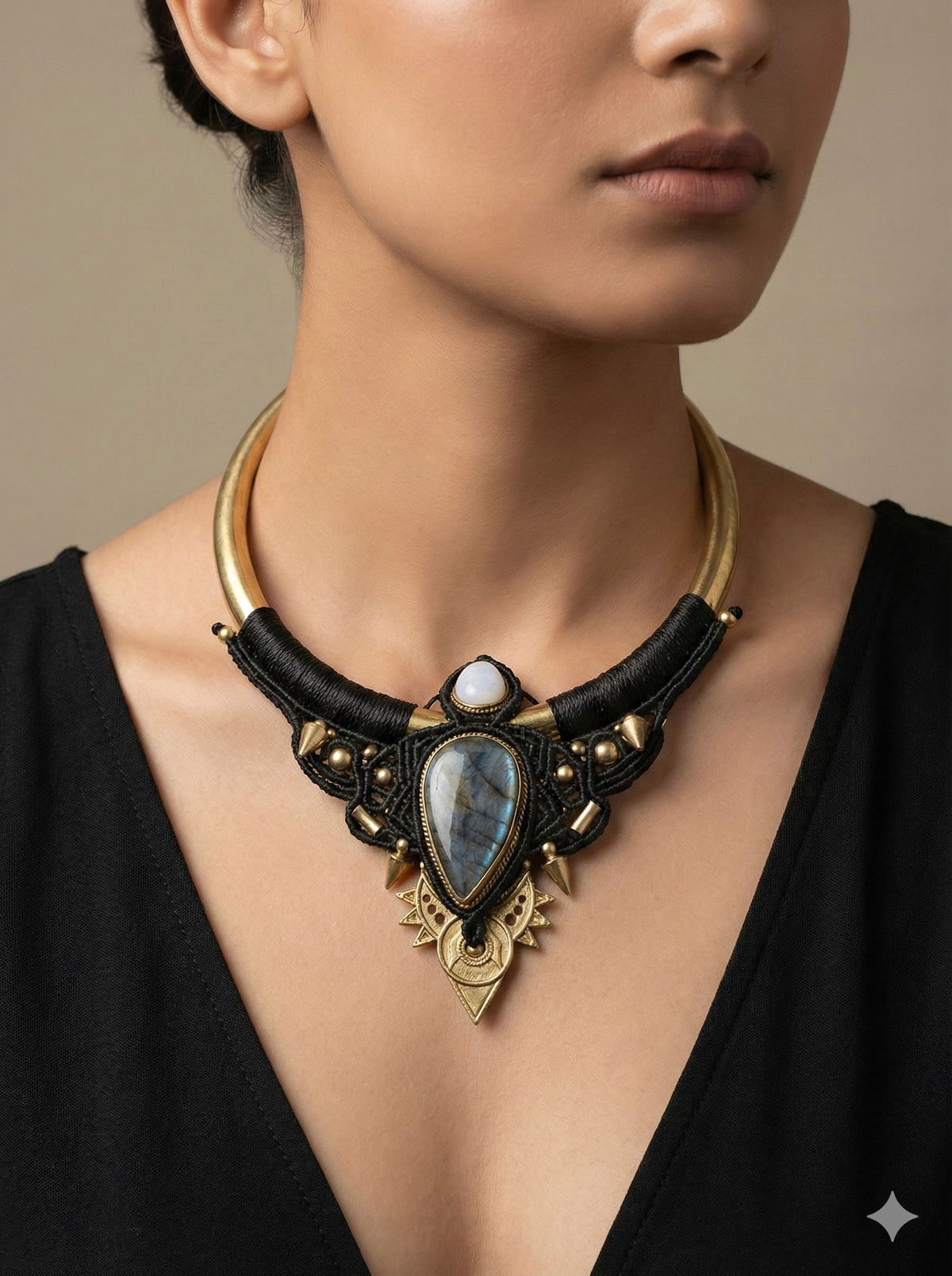 A woman wearing a black V-neck top and the Elvara Necklace, an ornate, handmade macramé piece adorned with gold, black, and stone accents. The necklace features a large teardrop-shaped labradorite moonstone at its center.