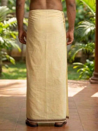 A person standing outdoors, shown from the back, is wearing the Mehfil Dhoti in light beige cotton with a simple border that adds an ethnic touch. Lush green plants and a tiled floor are visible in the background.