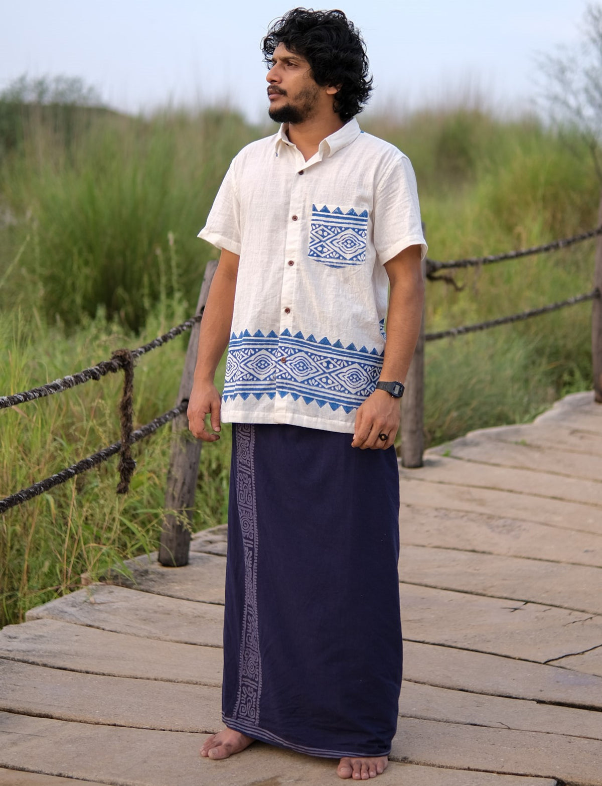 A man stands barefoot on a wooden pathway, dressed in the Bohemian Tribal Shirt—crafted from breathable cotton and adorned with blue block print patterns—and paired with a dark blue traditional wrap skirt. His outfit blends comfort with artisanal style, set against a backdrop of green grass and wooden railings.