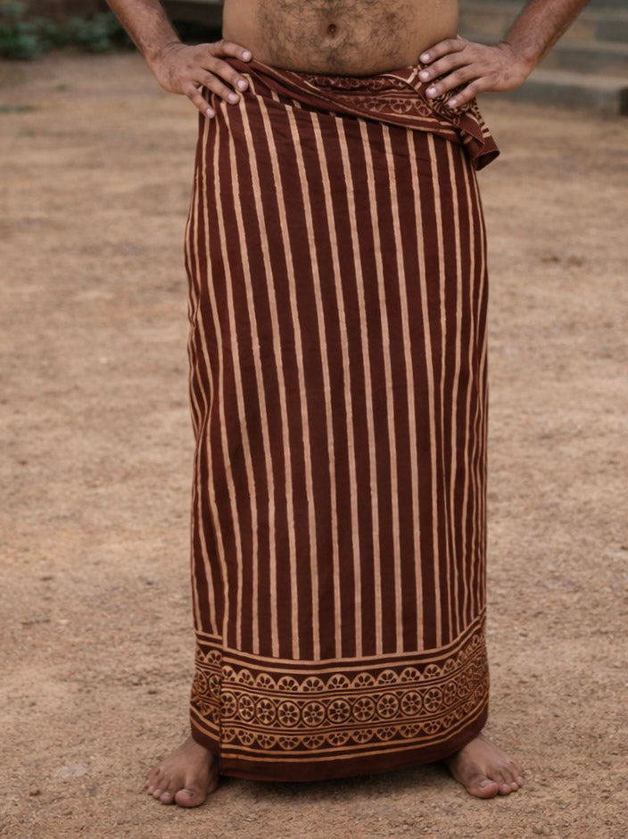 A person stands barefoot on dirt, wearing the brown Dhara Stripe Dhoti featuring vertical beige stripes and a patterned border. The upper body is not visible.