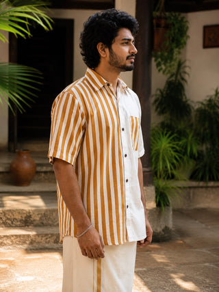 A man with curly hair stands sideways, wearing the Peetambar Shirt—a beige and brown kasavu-gold stripe short-sleeve shirt—paired with light pants. He is outdoors, surrounded by green plants and warm sunlight.