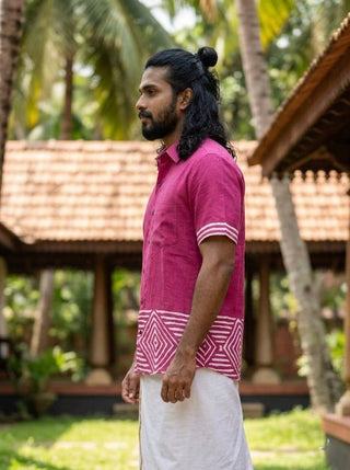 A man with long hair tied back wears the artisan-inspired, geometric block-print Raktara Shirt in pink paired with a white dhoti, standing outdoors against a backdrop of traditional tiled-roof buildings and palm trees.