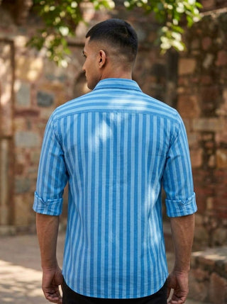 A man with short hair stands outdoors, his back to the camera, wearing the Neelpushpa Shirt—a long-sleeved shirt featuring blue and white vertical stripes and rolled-up sleeves. Behind him, a stone wall and greenery are visible.