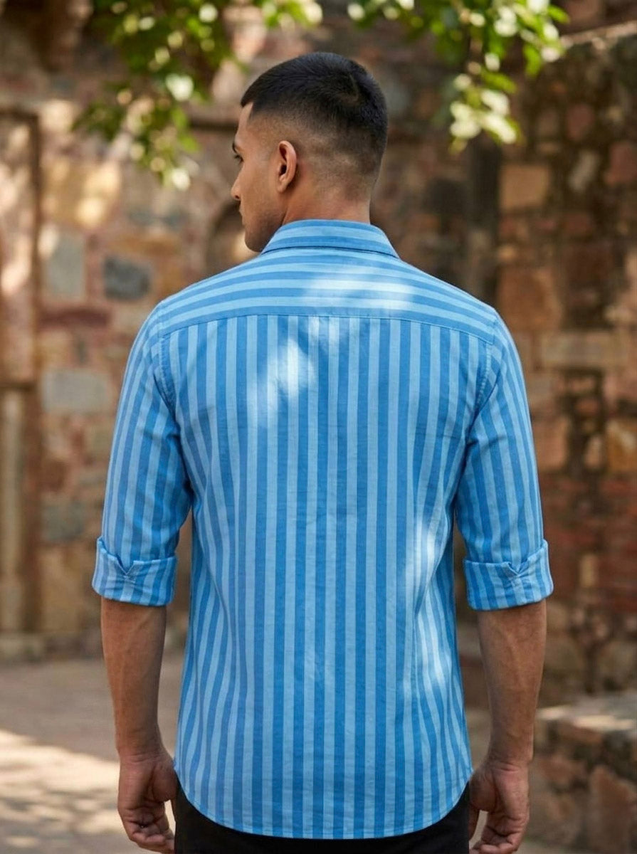 A man with short hair stands outdoors, his back to the camera, wearing the Neelpushpa Shirt—a long-sleeved shirt featuring blue and white vertical stripes and rolled-up sleeves. Behind him, a stone wall and greenery are visible.