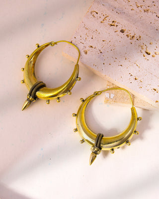 A pair of crescent-shaped Wisdom Earrings in gold with small metal beads and coil accents lies elegantly on a light cream and textured beige stone surface, showcasing their tribal-inspired design with sophisticated pointed tips and circular hoops.