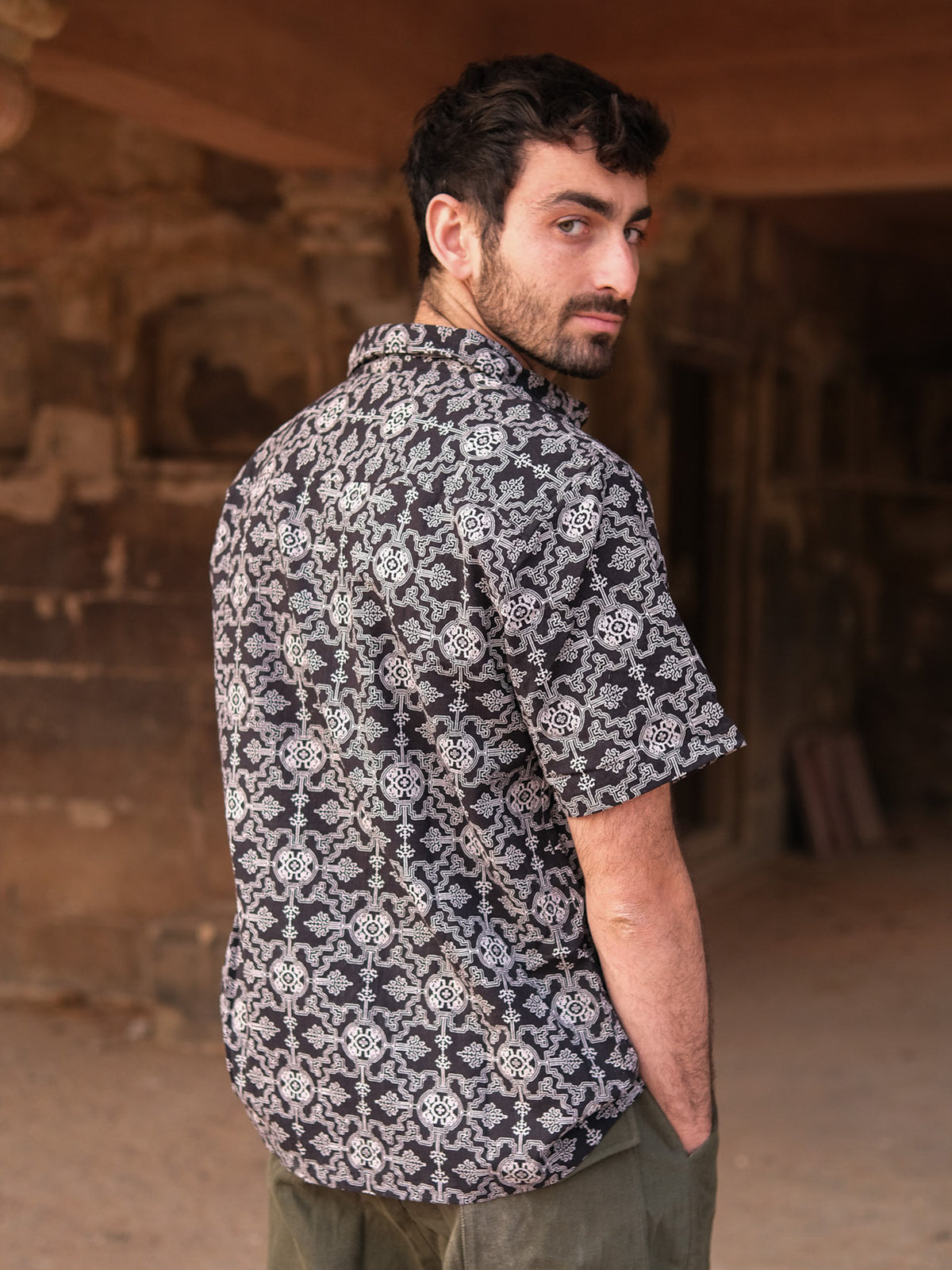 A man with short dark hair wears a black Shipibo Shirt with a patterned print, standing sideways and looking over his shoulder in a dim, rustic setting with stone walls and sandy ground.