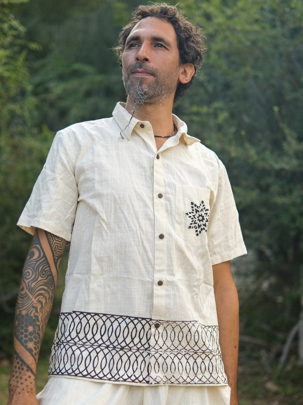 A man with a tattooed arm stands outdoors, thoughtfully gazing into the distance while wearing the Vairagya Shirt—a light-hued garment made from organic cotton and featuring striking geometric patterns. The soft blur of green foliage behind him highlights his mindful, eco-conscious fashion choice.