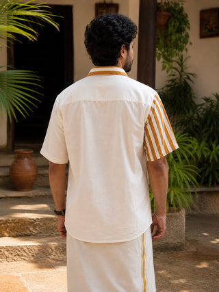 A man stands outdoors facing away, surrounded by plants and traditional clay pots, wearing the Peetambar Shirt—a white modern heritage piece with kasavu-gold stripe sleeves—and a white dhoti with a yellow stripe.
