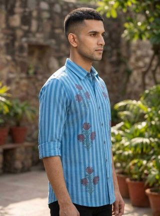A man stands outdoors in profile, wearing the Neelpushpa Shirt. The shirt features sky-blue stripes adorned with beautiful floral designs. Green plants in terracotta pots and a stone wall form the background.