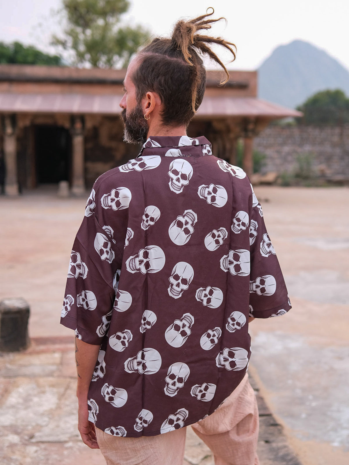 A man with dreadlocks stands outdoors, facing away. He wears the Shirova Shirt—a loose, dark top adorned with white skull patterns—paired with light-colored pants. A rustic building and mountains form the background.