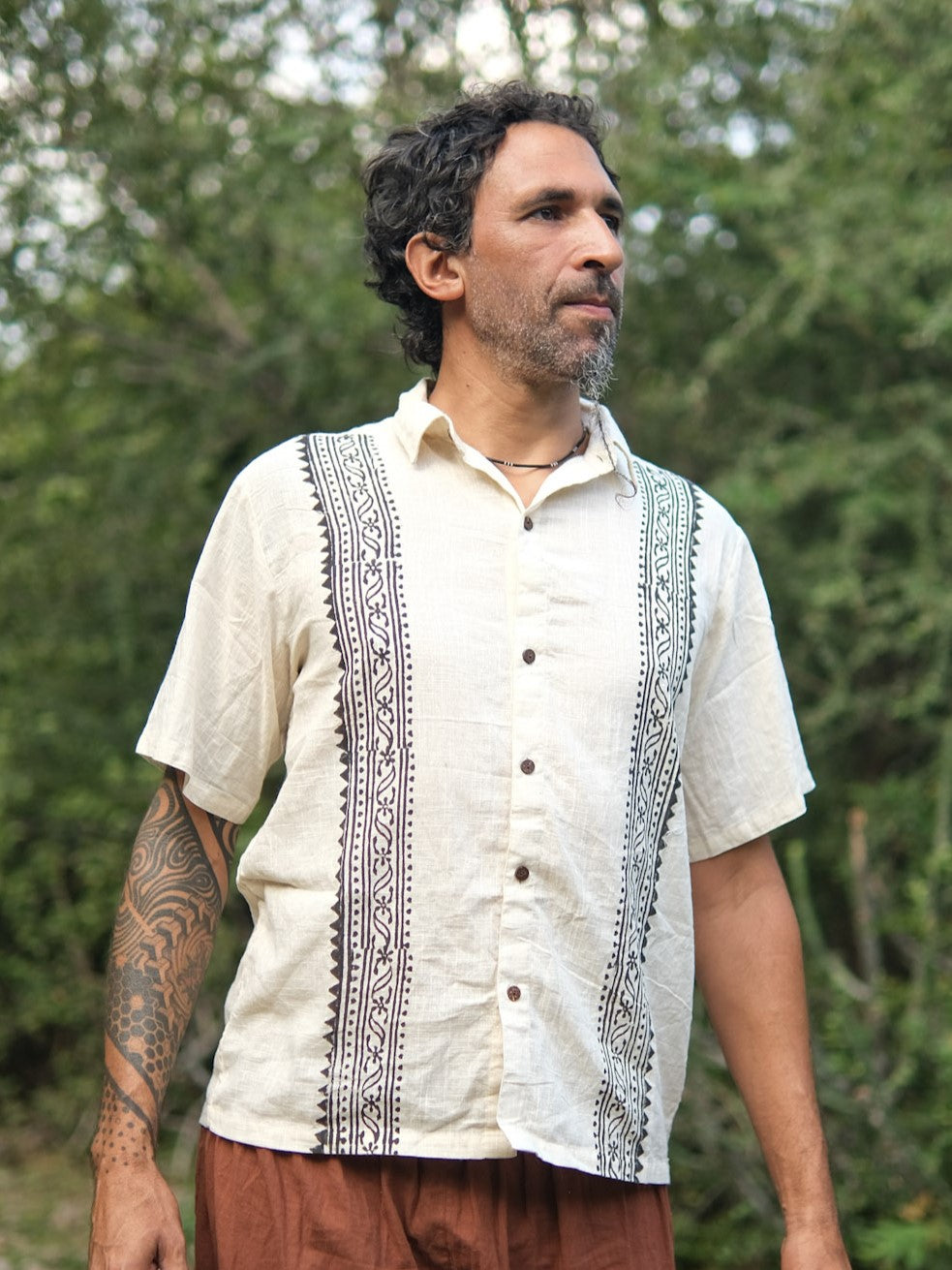 A man with curly hair and a beard stands outdoors, wearing the sustainable Gambheera Shirt Half-sleeve in cream, featuring intricate black patterns. Paired with brown pants, he displays tattoos on his right arm and gazes thoughtfully into the distance amid lush green foliage.