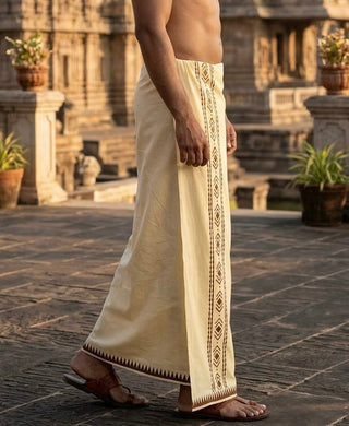 A man wearing the Mehfil Dhoti, crafted from cream-colored cotton and featuring patterned borders, stands outdoors on stone pavement, adding an ethnic touch against a backdrop of historic architecture and potted plants.