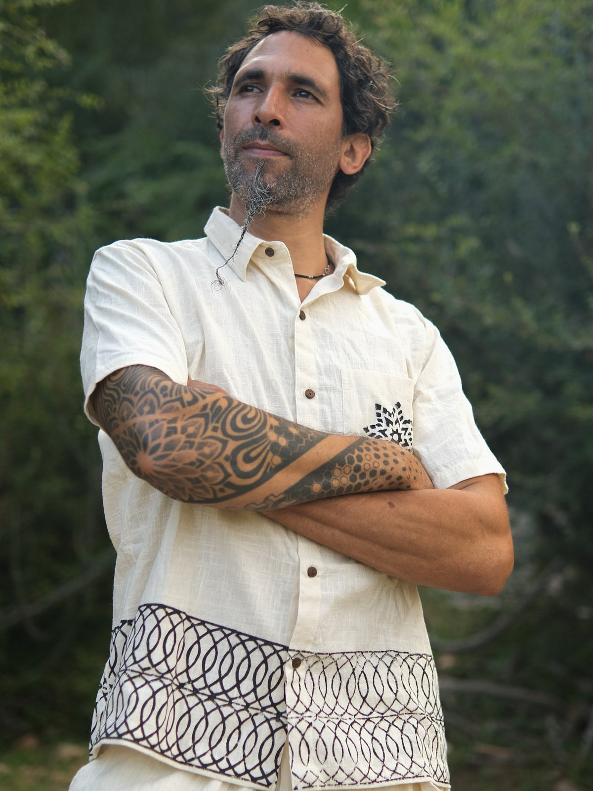 A man with curly hair and a patterned tattoo on his arm stands outdoors, arms crossed, thoughtfully gazing into the distance while wearing the Vairagya Shirt—a short-sleeved cream shirt crafted from organic cotton and adorned with decorative designs—perfectly embodying a sense of conscious fashion.