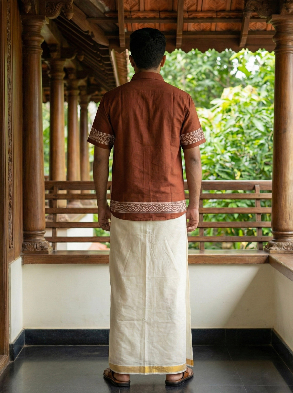 A man stands on a wooden veranda, facing away. He wears the Adhir Shirt—a brown, handcrafted-inspired cotton shirt with patterns—paired with a traditional white dhoti featuring a gold border. Lush greenery can be seen in the background, lending an ethnic contemporary charm to the scene.