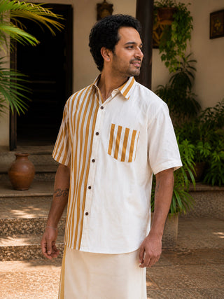 A man stands outside surrounded by lush green plants and rustic decor, smiling and looking to the side while wearing the Peetambar Shirt—a modern heritage piece in cream featuring bold brown vertical stripes on one side and on the pocket.