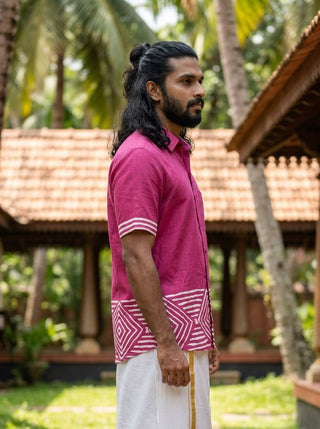 A man with long hair and a beard wears the Raktara Shirt—a berry-magenta geometric block-print shirt with white patterns—paired with a white dhoti. He stands sideways outdoors near traditional buildings and palm trees.