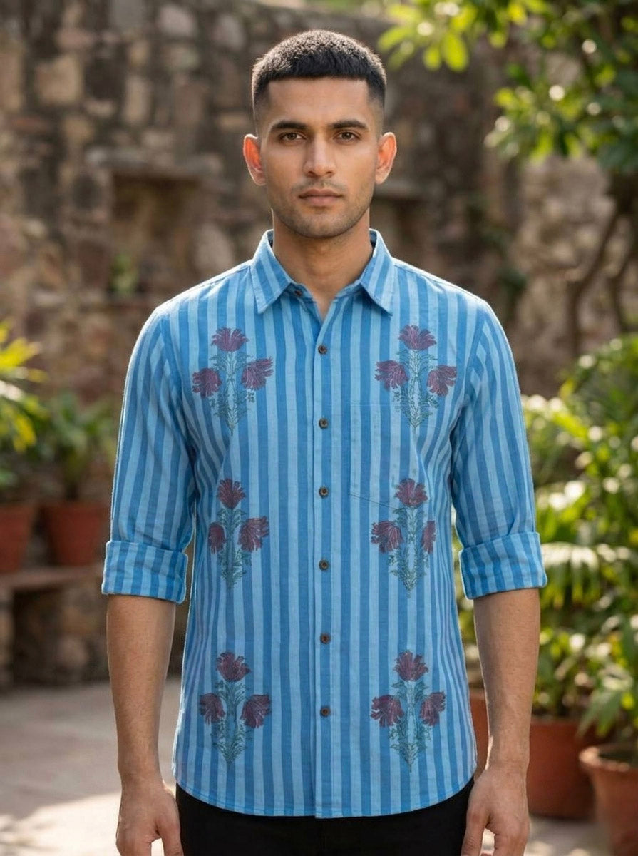A man stands outdoors wearing a blue Neelpushpa Shirt. He has short dark hair and a neutral expression, with green plants and a stone wall in the background.