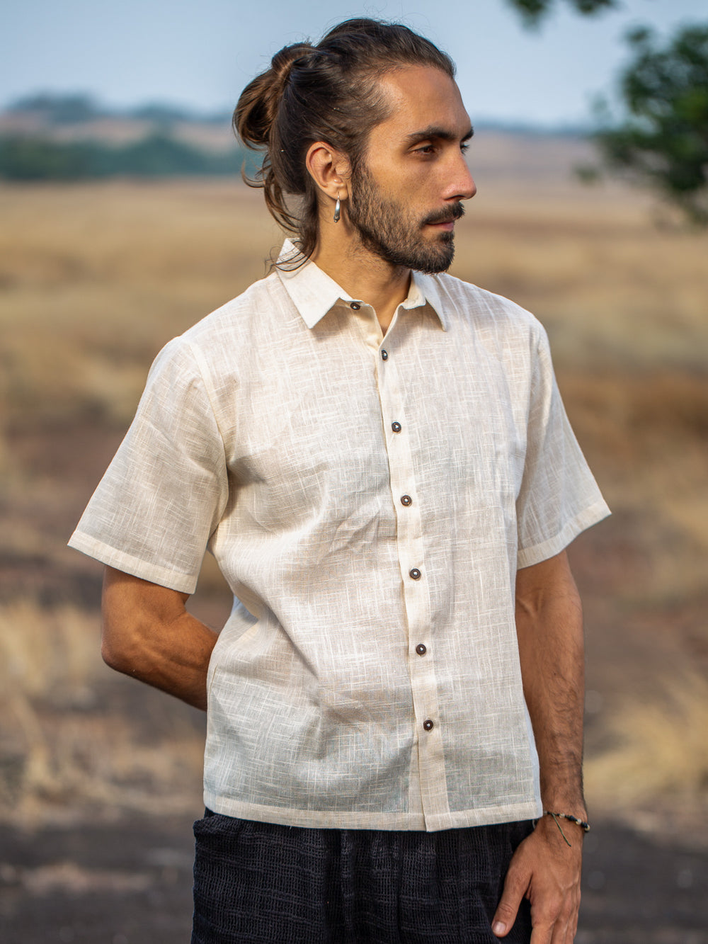 A man with long hair in a bun stands outdoors in a dry, grassy landscape, looking to the side with one hand behind his back, wearing the Yantra Shirt in Natural.