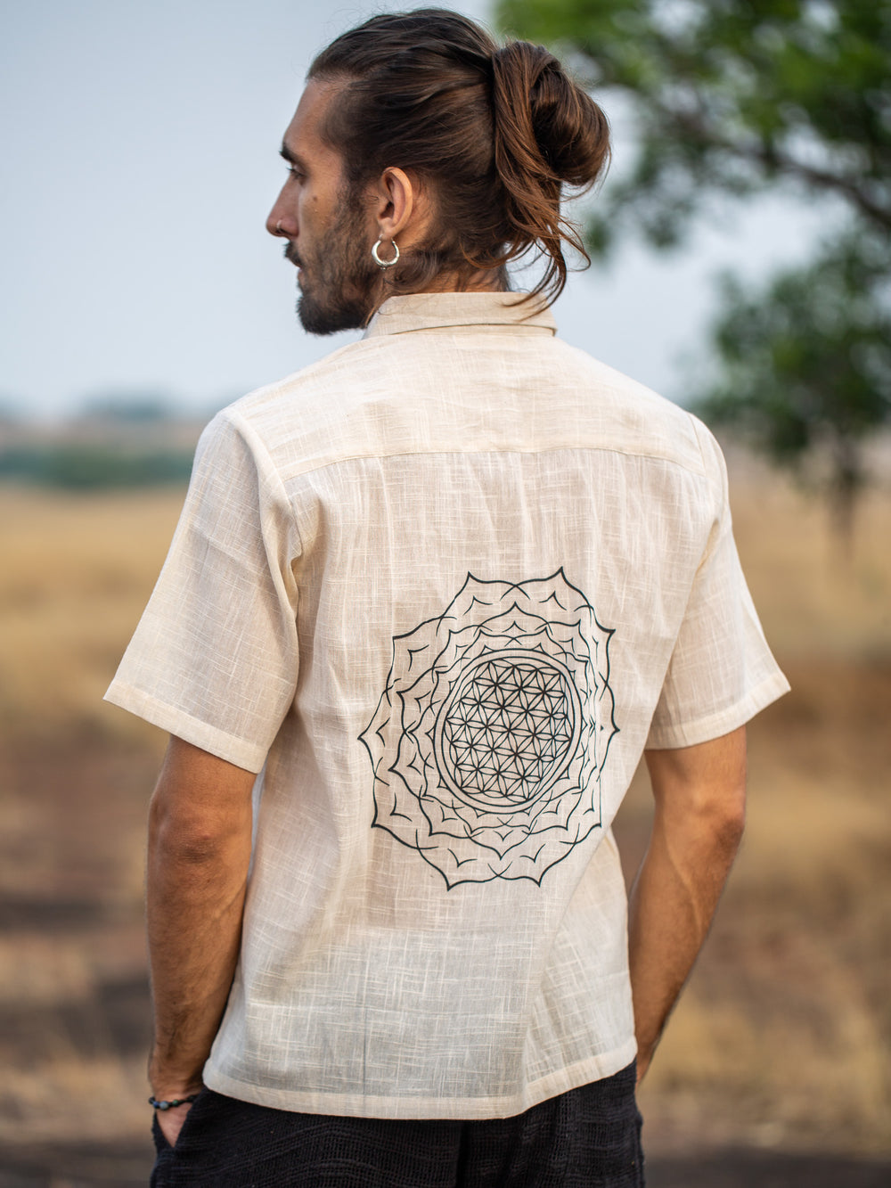 Wearing the Yantra Shirt in Natural, a person with long hair styled in a bun stands outdoors near dry grass and a tree. The light, breathable cotton shirt features a geometric mandala design on the back.